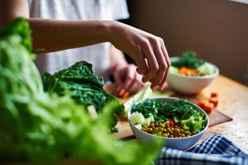 Hand pours green onions in a bowl with green peas, cucumbers, carrots, lettuce and dill standing on a table